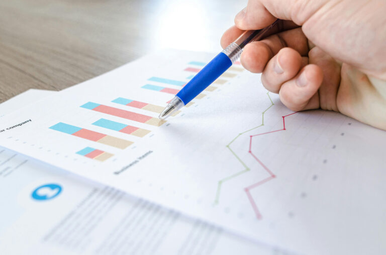 Hand holding a pen reviewing papers of financial graphs.